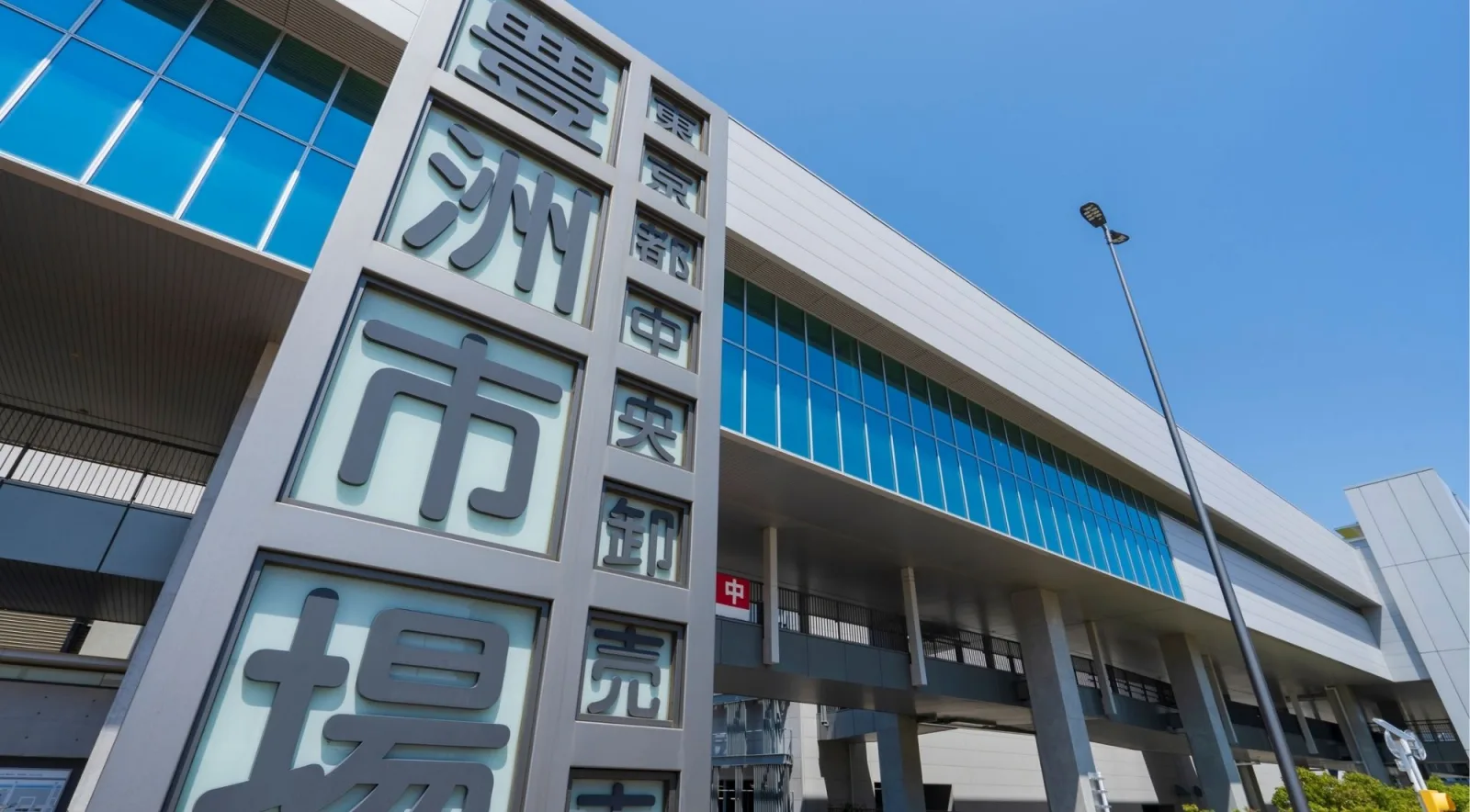 Toyosu Market entrance sign reading Tokyo Metropolitan Central Wholesale Market against the modern glass building facade